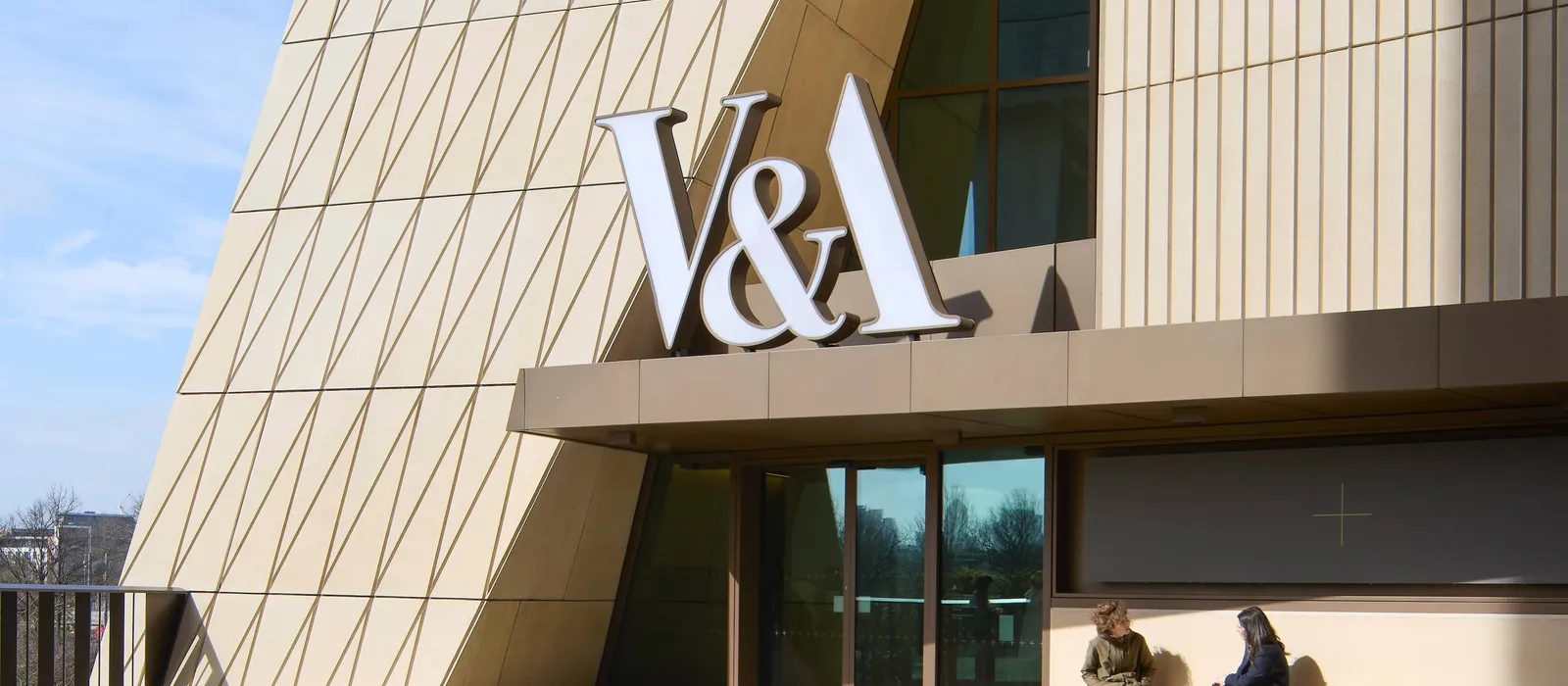 Exterior view of the V&A East building, featuring a striking cream-coloured façade with geometric diagonal paneling and a large “V&A” sign above the entrance. Two people sit and talk on a bench beside the glass doors, while sunlight casts strong shadows a