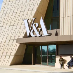 Exterior view of the V&A East building, featuring a striking cream-coloured façade with geometric diagonal paneling and a large “V&A  sign above the entrance. Two people sit and talk on a bench beside the glass doors.