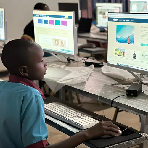 A child using a desktop computer in a classroom, with other students working in the background.