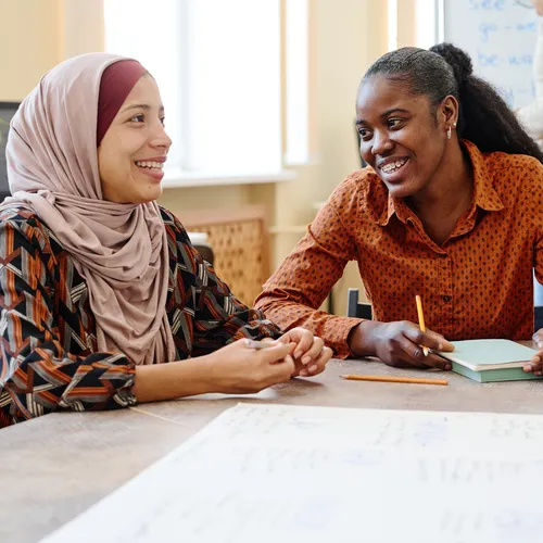 A group of people walking away from the camera on a paved path, with a person in a bright blue winter coat carrying a small child in the foreground. Grey cloudy sky overhead. The scene suggests people in transit or on a journey.