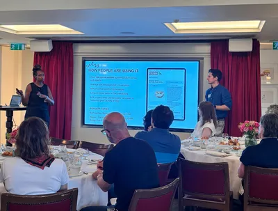 Ben and Trish present at the front of a bright conference room. Ben stands beside a large screen displaying a London Museum slide about how people are using an AI-powered collections experience, while Trish speaks to the seated audience. Attendees sit around round tables set for breakfast, listening attentively.