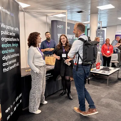Emma and Cassie chatting with a delegate. The stand features a banner about partnering with public sector organisations to design and deliver digital services, with other exhibitors visible in the background.