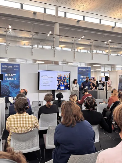 Dan speaking from the Impact Stage. He gestures expressively while presenting alongside a slide titled ‘Where we’re headed,’ as the audience listens with headsets.