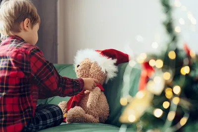 Young child in festive pyjamas sitting on a sofa, dressing a teddy bear in a Santa hat and scarf, with a decorated Christmas tree in the foreground.