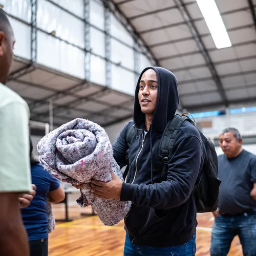 Two women embracing outdoors, one wearing a pink head covering appears to be a cancer patient, while the other woman is kissing her cheek. Both are smiling warmly in a sunlit park or garden setting.
