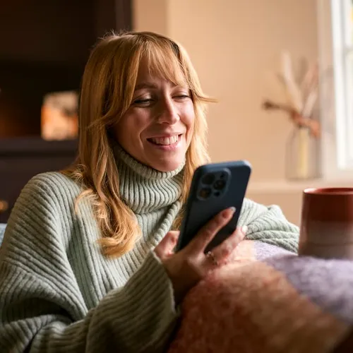 A person sitting on a sofa at home, smiling and looking at their phone.