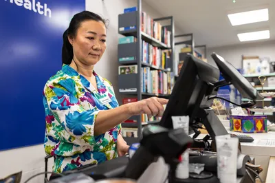 A charity shop volunteer in a colourful floral top uses a touchscreen till at the shop counter, with shelves and displays in the background.