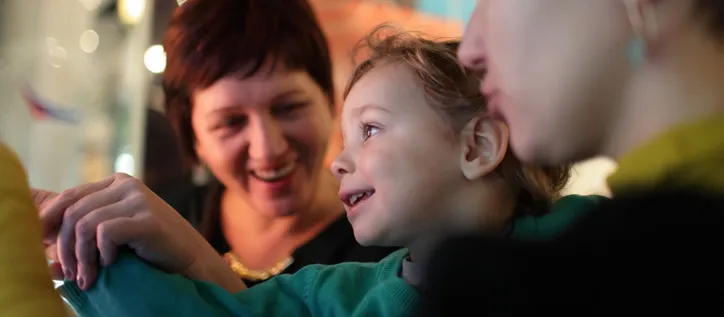 A young child smiles and reaches towards an exhibit while two adults look on during a museum or gallery visit.