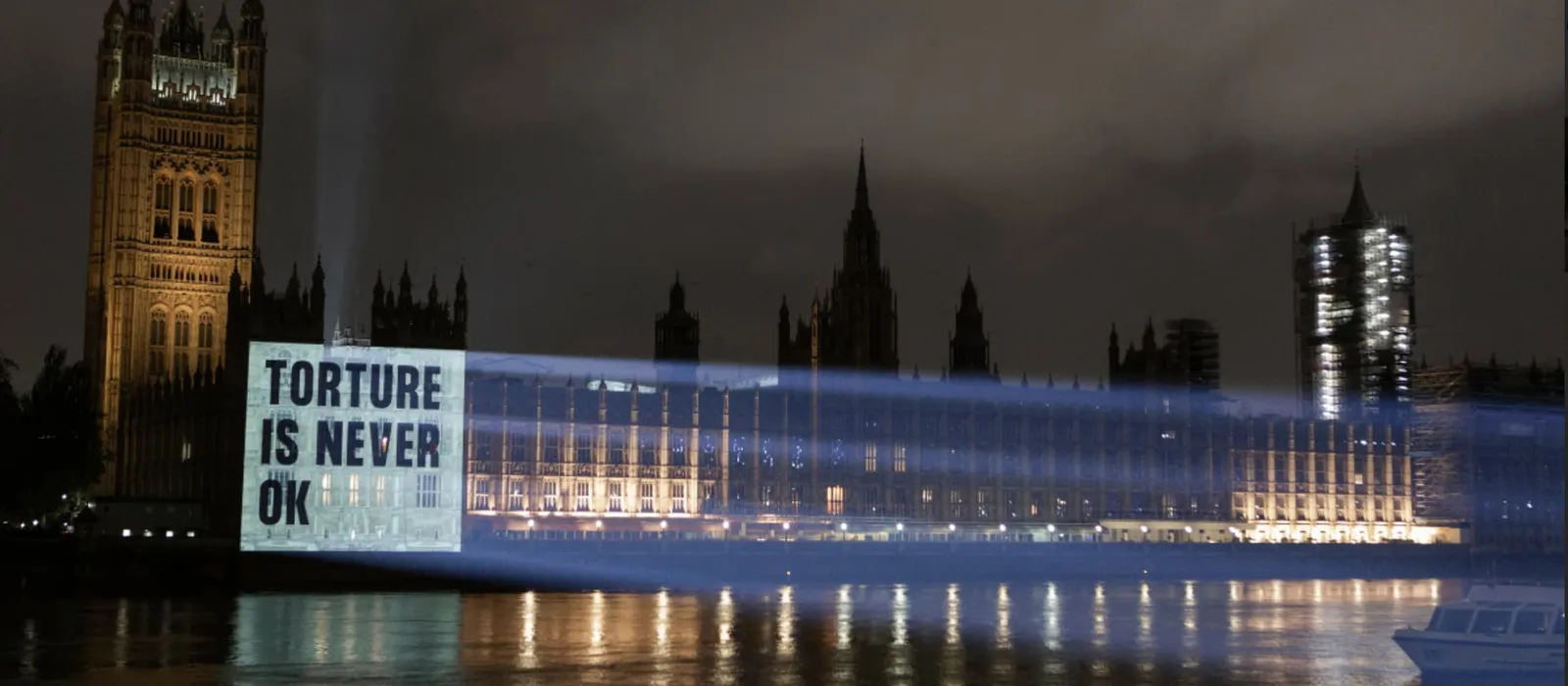 Night-time view of the Palace of Westminster beside the River Thames, with a bright projection cast onto the side of the building that reads "Torture is never okay".