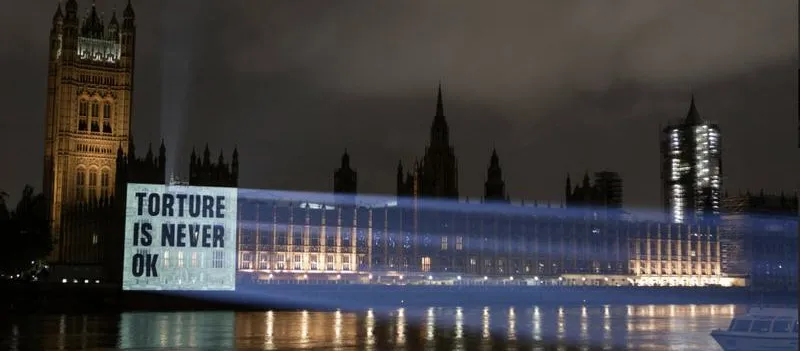 Night-time view of the Palace of Westminster beside the River Thames, with a bright projection cast onto the side of the building that reads "Torture is never okay".