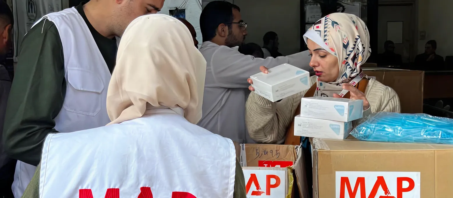 Three aid workers wearing white vests labeled “MAP – Medical Aid for Palestinians” organise boxes of supplies.