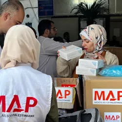Three aid workers wearing white vests labeled “MAP – Medical Aid for Palestinians” organise boxes of supplies.
