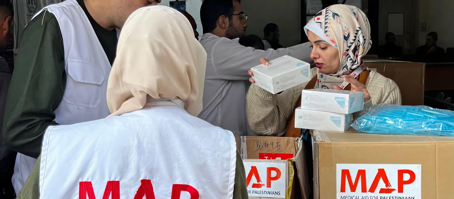 Three aid workers wearing white vests labeled “MAP – Medical Aid for Palestinians” organise boxes of supplies.