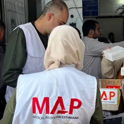 Three aid workers wearing white vests labeled “MAP – Medical Aid for Palestinians” organise boxes of supplies.
