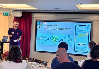 Matt from Migration Museum stands at the front of the room presenting to attendees.Behind him, a large screen illustrates an AI concept that connects museum collections to visitors' personal migration stories through user input, AI analysis and related objects.