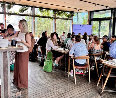 Attendees at a charity leader breakfast event mingle and chat around tables in a bright, plant-filled venue at The Lighterman in King’s Cross. One guest collects food at the buffet while others are seated in conversation. A screen in the background displays a slide from the Charity Digital Skills Report.