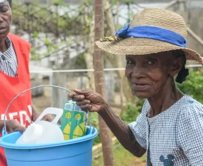 An older woman wearing a straw hat with a blue ribbon smiles while carrying a blue bucket containing hygiene supplies. Another person wearing a red vest stands beside her outdoors in a green, rural setting.